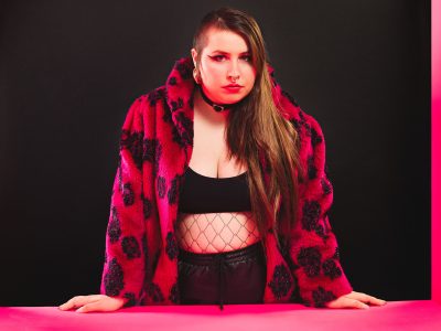 Sängerin Finna in roter Felljacke mit schwarzem Muster, schwarzem Halsband, Top und Hose stützt sich mit den Händen auf einem pinken Rahmen vor schwarzem Hintergrund ab.