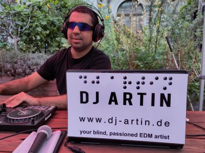 DJ Artin sitzt mit Sonnenbrille und Kopfhörern an den Turntables, vor ihm ein Schild mit der Aufschrift: DJ Artin, www.dj-artin.de; your blind, passioned EDM artist
