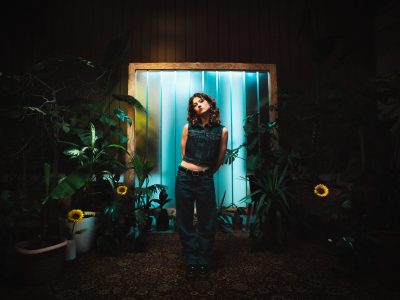 Luana trägt Jeans-Oberteil und-Hose und steht vor einer beleuchteten Bretterwand, umgeben von Grünpflanzen und Sonnenblumen.
