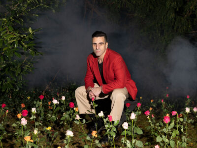 Der Musiker sitzt in einem roten Bläser lässig in einer Wiese mit Blumen.